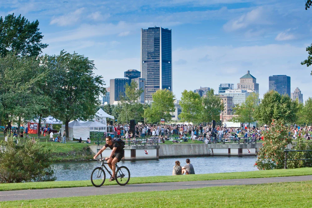 People enjoying a sunny day at a riverside park in Saint-Henri, with Montreal’s skyline visible in the background.