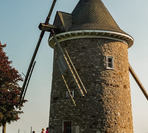 Historic stone windmill in Pointe-Claire, Montreal, with wooden blades and a conical roof, photographed on a sunny day with people walking nearby.